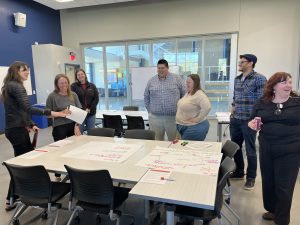 group of people at a meeting, working together in discussion. There is a table with large paper in front of them, with writing on the paper. 